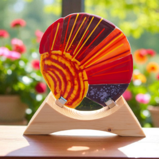 Handmade fused glass sunrise ornament in red, orange, and yellow with a natural wooden stand, displayed in sunlight with flowers in the background.