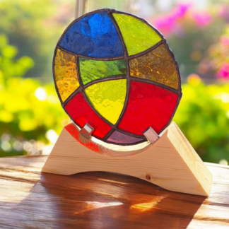 Round stained glass panel with red, yellow, blue, green, purple, and orange sections, mounted on a pale wooden stand and lit by sunlight from a window.