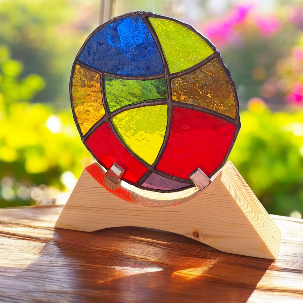 Round stained glass panel with red, yellow, blue, green, purple, and orange sections, mounted on a pale wooden stand and lit by sunlight from a window.