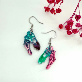 A pair of lightweight resin earrings shaped like dinosaur skulls in swirling pastel rainbow hues of pink, teal and purple, displayed on a textured background.