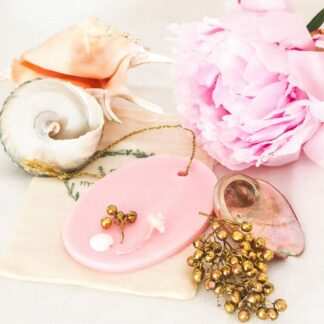 Pink oval shaped soy wax tablet with gold hanging loop, decorated with gold, white and pink botanicals. The wax melt/room freshener is on a white background resting on a muslin bag, surrounded by a large pink peony, gold berries, mother of pearl and large sea shells which give an air of relaxation and luxury.