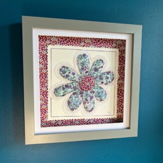 A box frame hanging on a blue wall featuring an appliqued blue and pink daisy in floral fabric