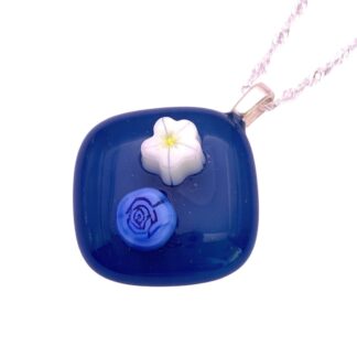A square, glossy blue glass pendant with a white flower and a blue rose design. It hangs elegantly from a delicate silver chain, exuding a serene charm.