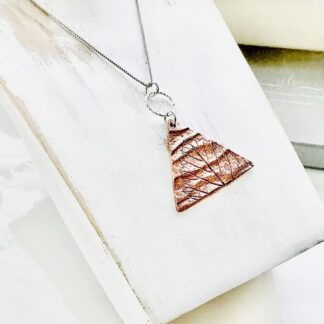A handmadectriangle shaped copper clay pendant that has been textured by pressing a leaf into the clay, it has a sterling silver ring through the silver jump ring and a sterling silver chain. It is displayed on a white washed wood pendant stand.