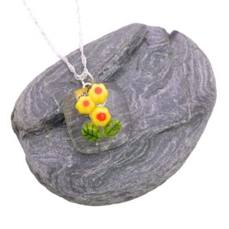 Pendant featuring yellow daffodils with red centers, encased in clear glass, on a silver chain. Set against a textured gray rock background.