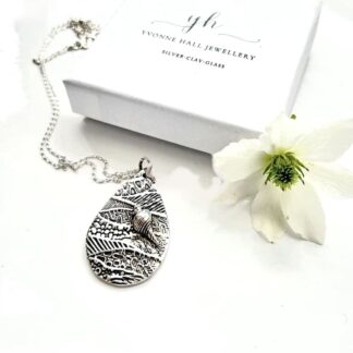 A fine silver 999 pendant, tear drop shape which is textured to resemble a beach with a small sea shell, it has a sterling silver chain. It is displayed a white background with a white flower.