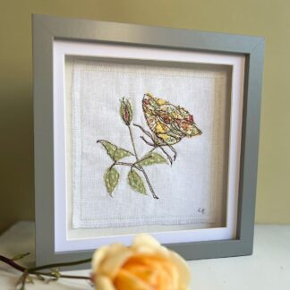 Textile art of a golden yellow single stem rose in full bloom with a single bud. The grey frame is standing on a shelf with a yellow rose laying in front.