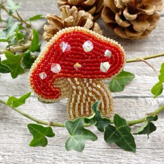 Cottagecore-style hand beaded mushroom pin in red, white, and earthy tones, by DewCatDesigns, photographed in natural light.