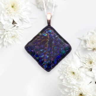 A multicolored, iridescent dichroic glass pendant with a textured surface hangs from a silver plated chain, surrounded by delicate white chrysanthemums on a soft background.