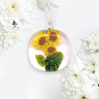 A delicate glass pendant featuring bright yellow sunflowers with green leaves, set against a soft pink backdrop, is surrounded by white chrysanthemums.