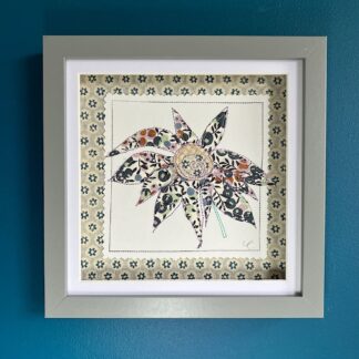 A grey box frame on a blue wall with an appliqued daisy flower in pink and green patterned fabric