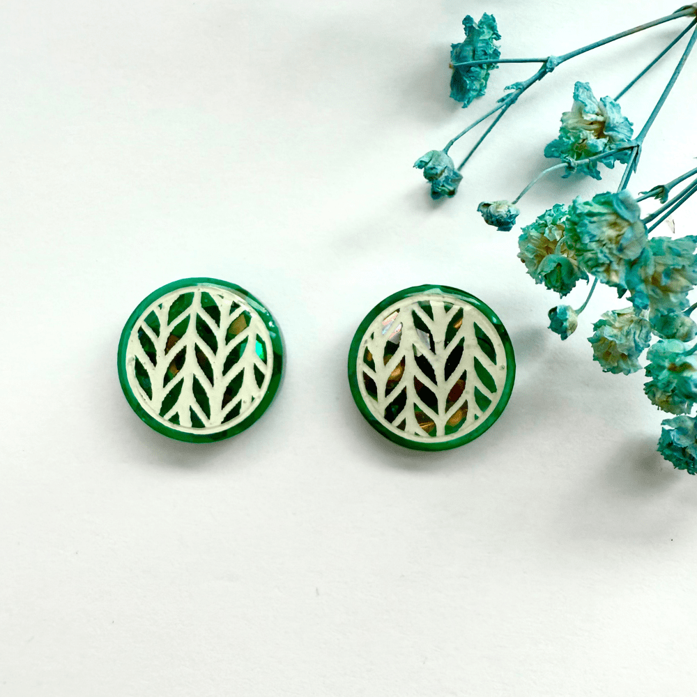 Pair of teal knitted-effect stud earrings with holographic glitter background lying flat beside dried blue flowers.