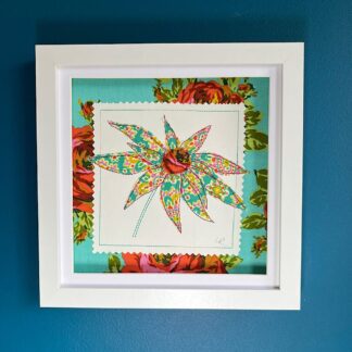 A white box frame hanging on a blue wall featuring a bright appliqued flower with rose printed fabric