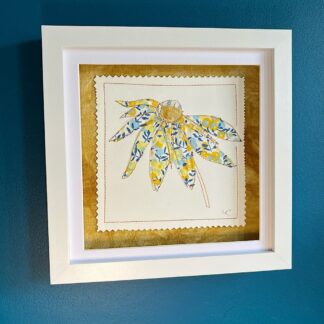 A white box frame on a blue wall with a gold and blue appliqued flower on a white background