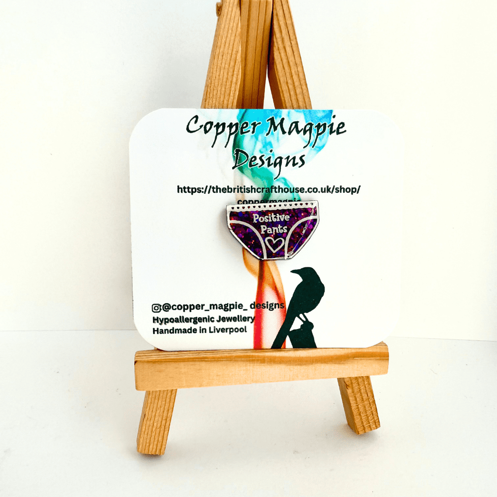 Pink glitter colourshift positive pants pin badge on branded Copper Magpie Designs card resting on a minature easel