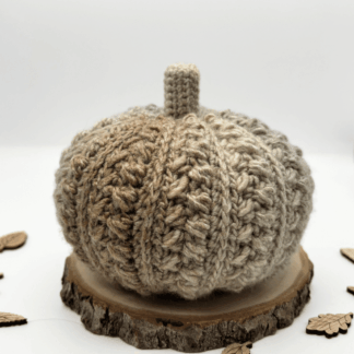 the photo shows a crochet pumpkin in shades of beige and light grey. It is textured and is resting on a log slice, there are small wooden leaves scattered around