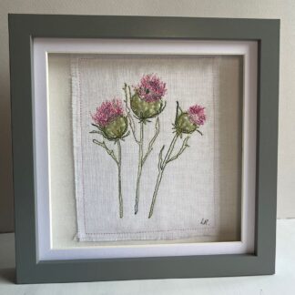 Framed embroidery artwork of three pink thistles, single stem side by side in a grey frame.