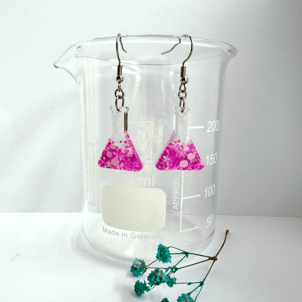 Conical flask shaped earrings with white bubble detail, half filled with candy pink glitter with stars hang from surgical steel ear hooks from a lab beaker.