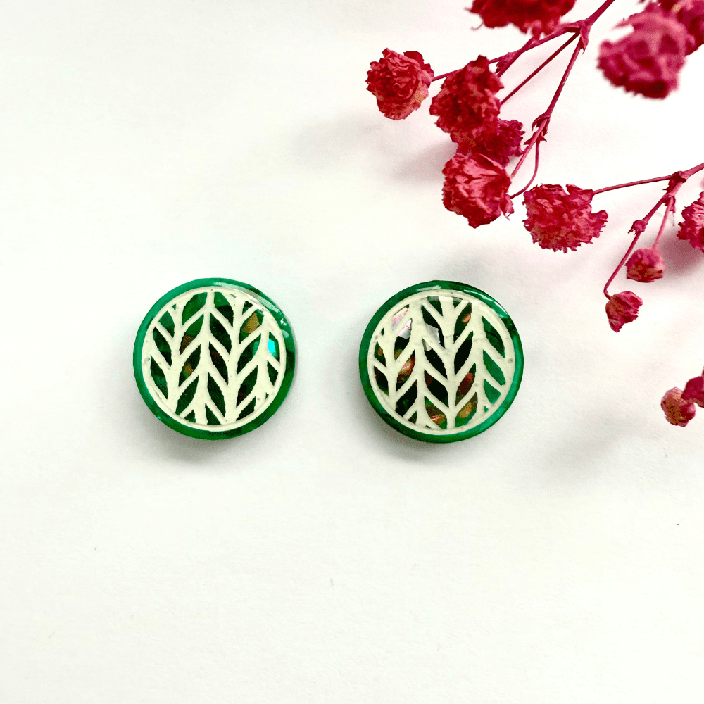 Pair of teal knitted-effect stud earrings with holographic glitter background lying flat beside dried pink flowers.