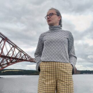 Woman dressed in grey turtlenck and yellow check trousers standing in front of Forth Bridge