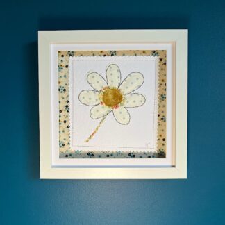 A white box frame on a blue wall featuring an applique of a single daisy in cream and grey spot fabric. Background white card surrounded with blue floral fabric.