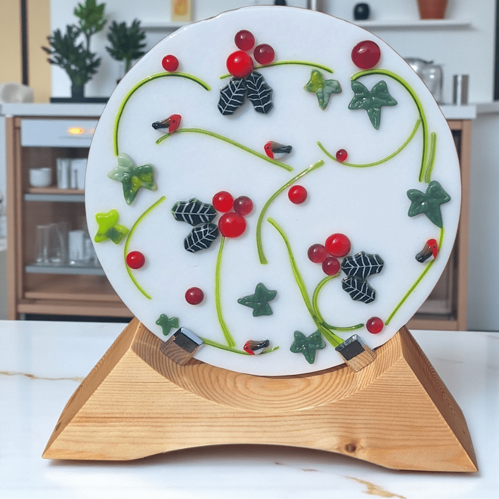 Handmade fused glass Christmas decoration featuring holly and ivy leaves, glossy red berries, and tiny robins, all set against a smooth white glass base. Displayed on a natural wooden stand, this festive glass ornament makes a unique seasonal centrepiece or mantel display. A perfect Christmas gift or keepsake for nature lovers, combining glass art craftsmanship with traditional holiday charm.