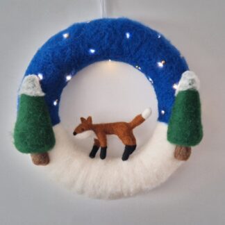 Fox on snowy wreath surrounded by two green trees, dark blue sky with fairy lights as stars