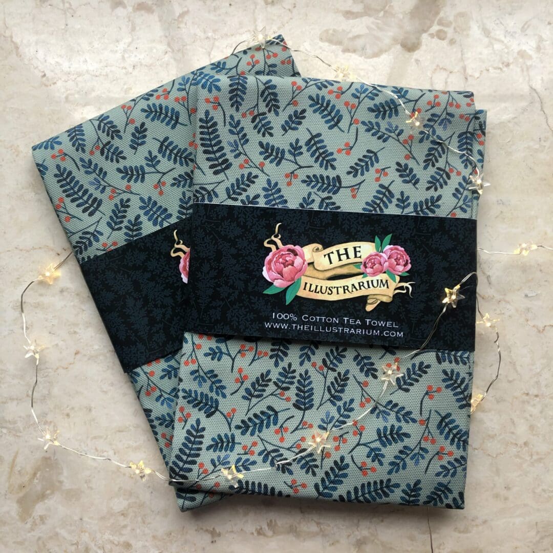Two folded Berries Tea Towels with The Illustrarium’s floral and berry design packaging sleeve, surrounded by twinkling fairy lights.