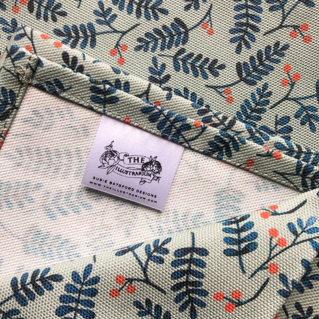 Close-up of the Berries Tea Towel label with The Illustrarium logo and stitched seam showing the textured cotton weave.