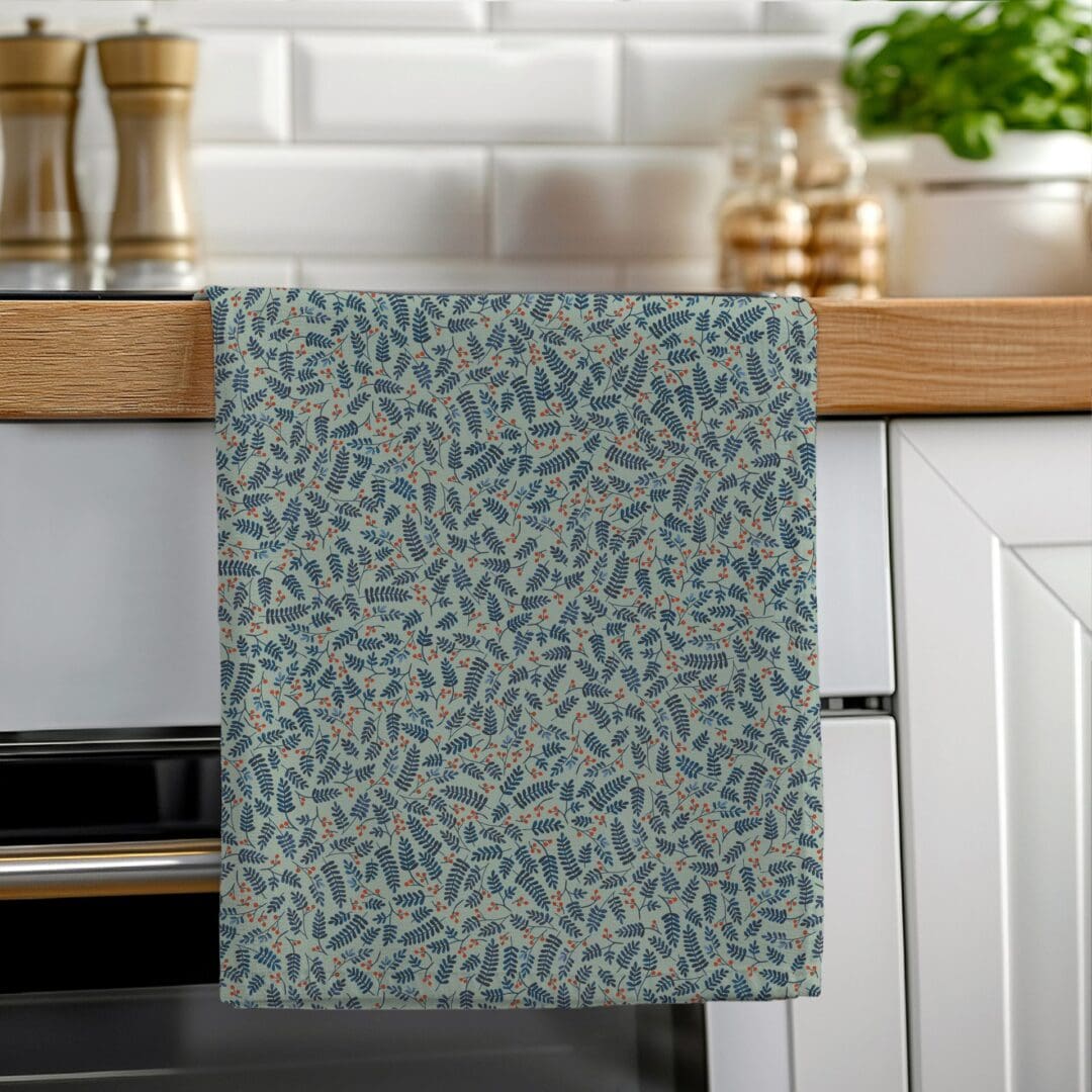 A soft green cotton tea towel featuring a hand-illustrated botanical berries pattern hanging neatly on a wooden oven rail in a bright kitchen.