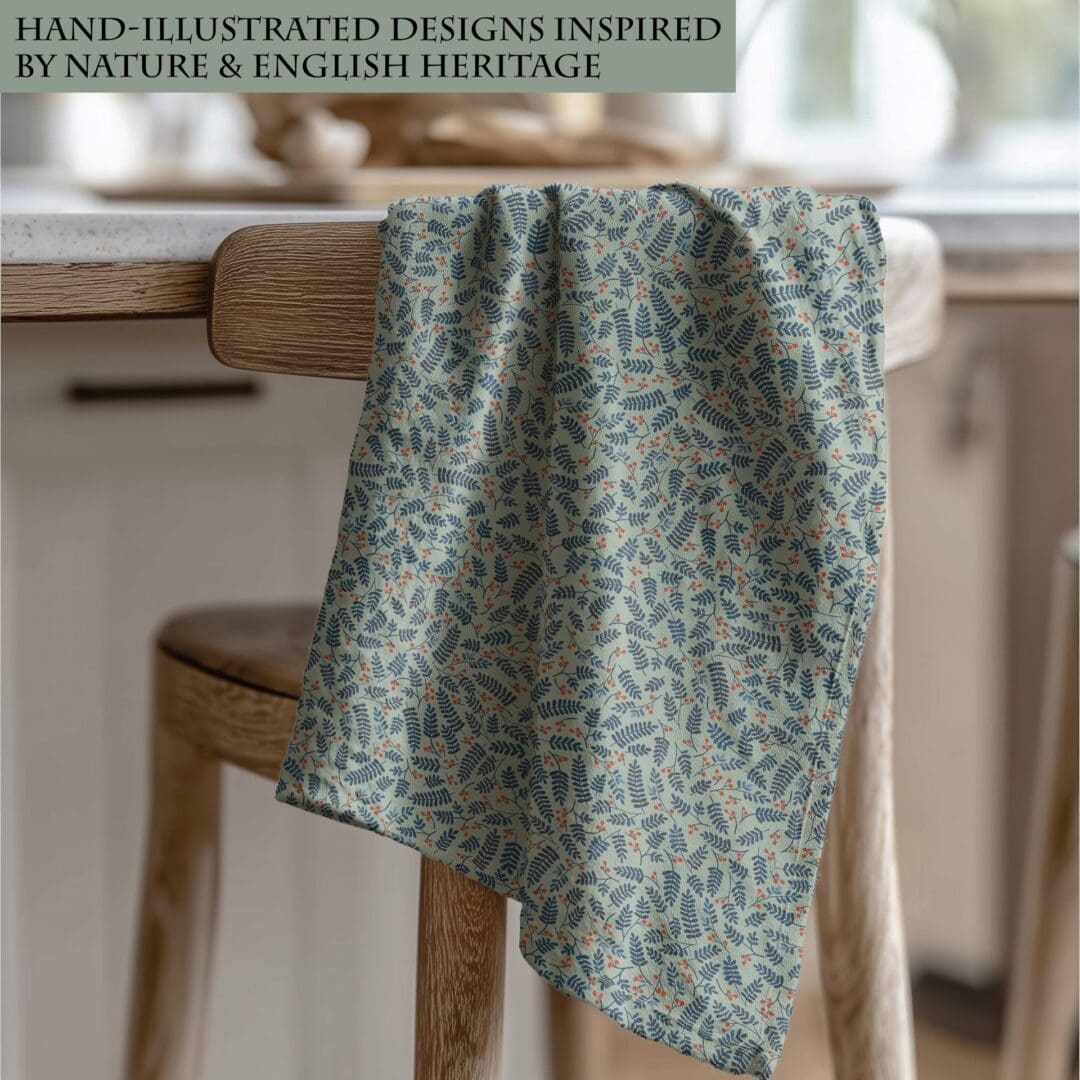 Botanical Berries cotton tea towel draped over a wooden chair in a cosy kitchen, featuring blue foliage and red berry illustrations on a soft green background.