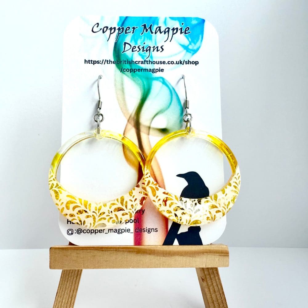 Yellow hoop earrings with lace and leaf detail hang from surgical steel ear hooks on a branded Copper Magpie Designs jewellery card.