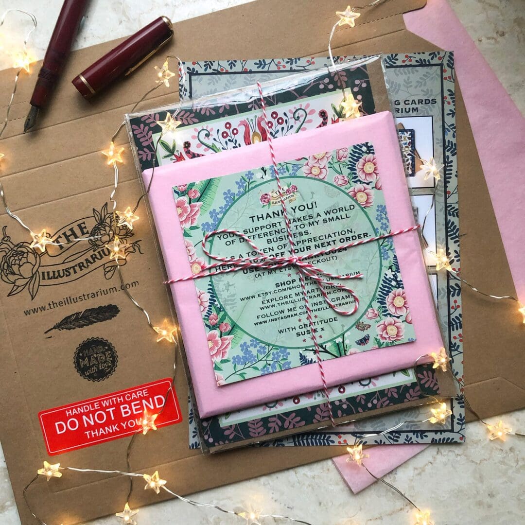 A flatlay of The Illustrarium’s eco-friendly packaging, featuring a tied bundle of greeting cards, a mounted butterfly art print wrapped in tissue, and a branded envelope with flyer and voucher.