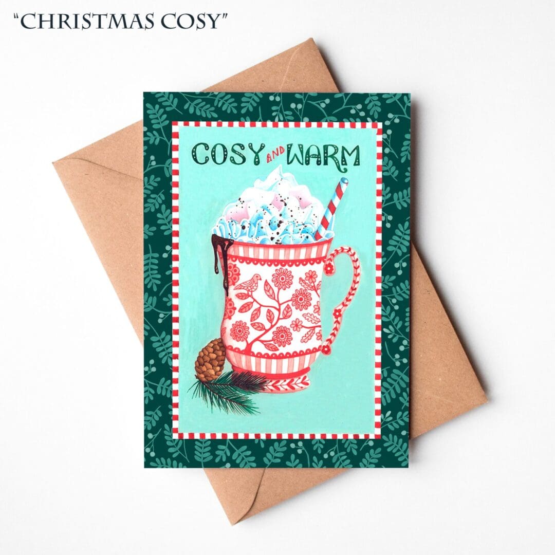 A hand-illustrated Christmas card featuring a steaming red mug of hot chocolate with the words “Cosy & Warm,” shown resting on a natural brown Kraft envelope.
