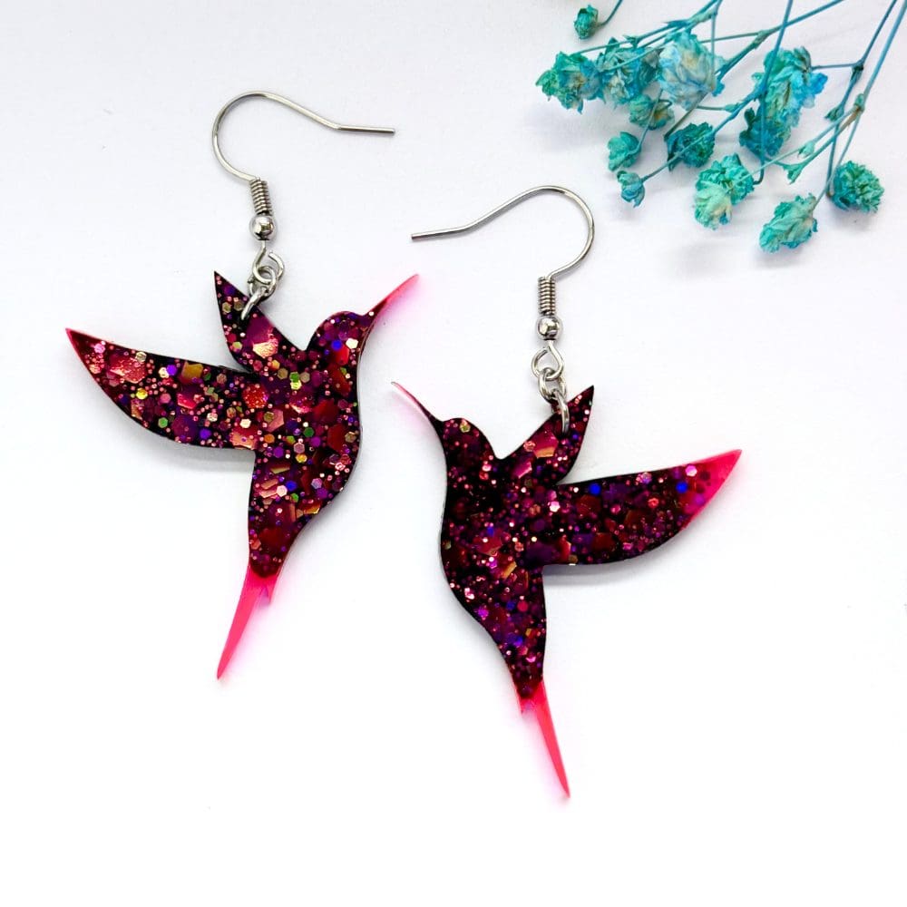 Glittering black hummingbird with pink tips earrings against a soft floral background. Made using hypoallergenic hooks