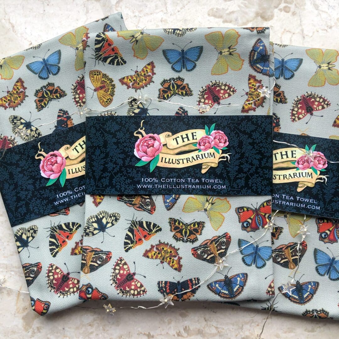 Three folded Folk Butterflies Tea Towels with The Illustrarium’s floral logo sleeve, surrounded by fairy lights, showing vibrant butterfly designs.