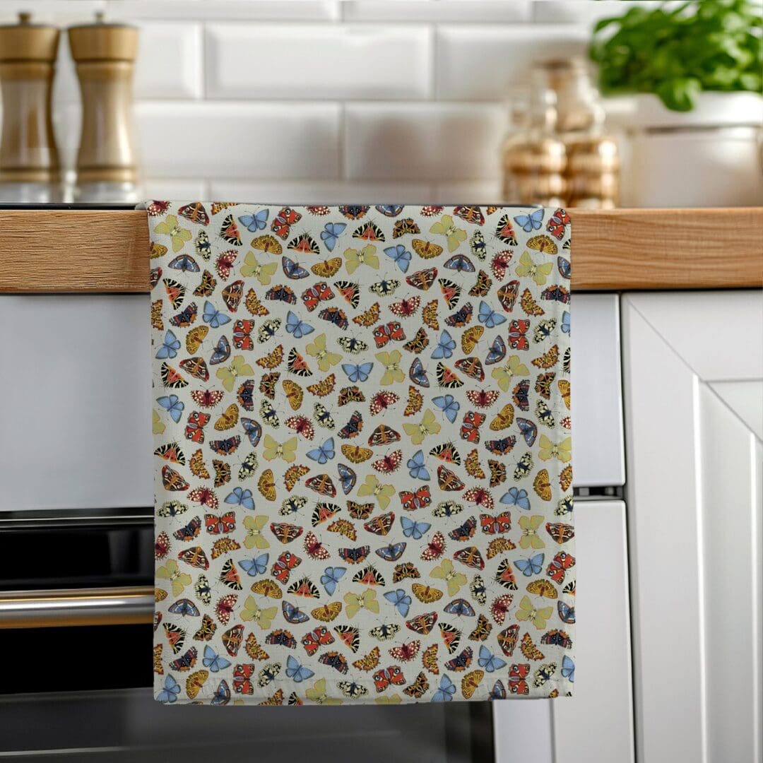 A colourful cotton tea towel featuring hand-illustrated butterflies in folk art style, hanging neatly on a wooden oven rail in a bright modern kitchen.