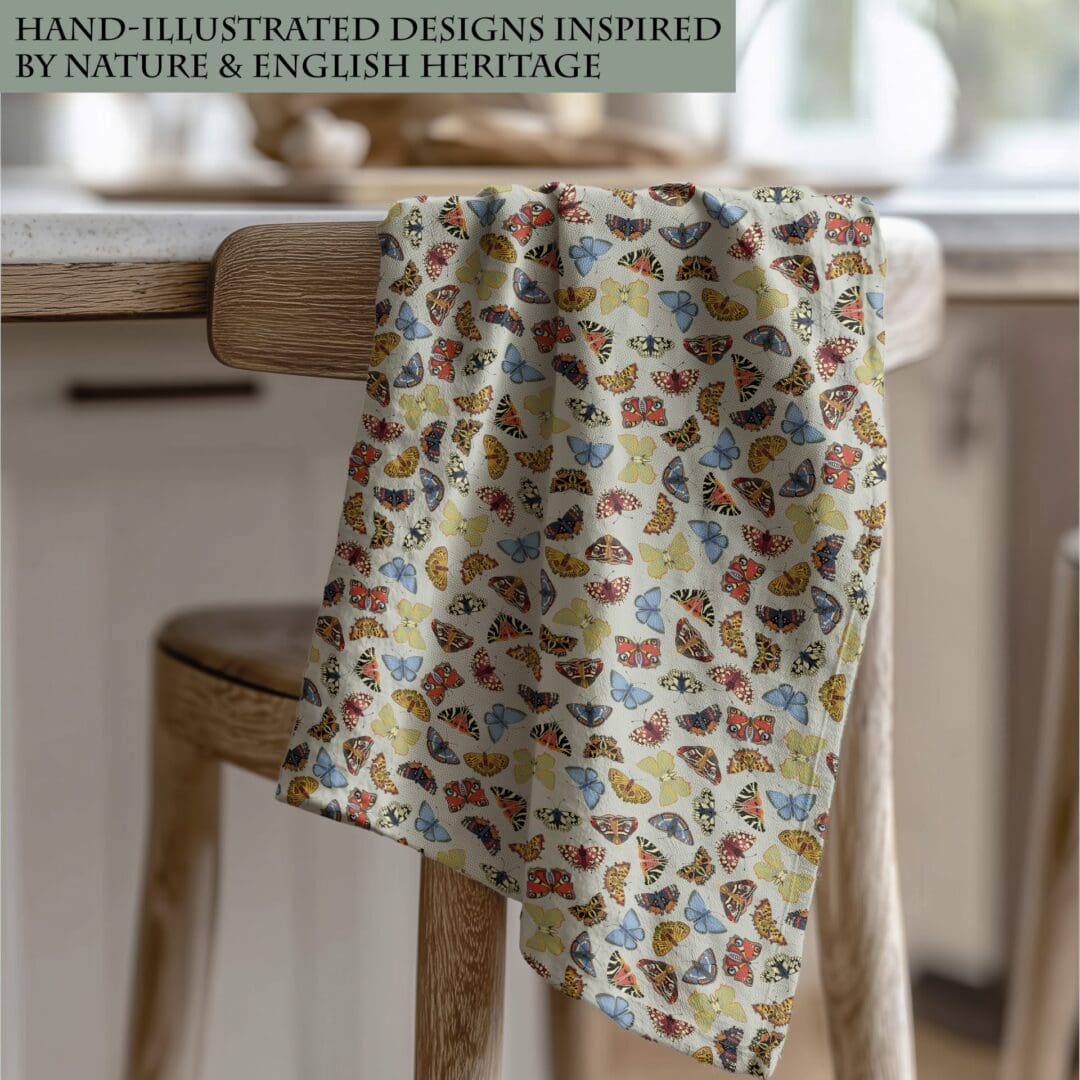 Folk Butterflies cotton tea towel draped over a wooden chair in a cosy kitchen, featuring colourful butterfly illustrations on a soft sage background.