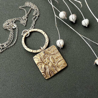 A handcrafted bronze clay double pendant, the bottom half is square with a textured surface, the top half is a bronze round ring with the same texture and they are linked together with a sterling silver jump ring. It has a sterling silver chain and is displayed on a dark grey surface.