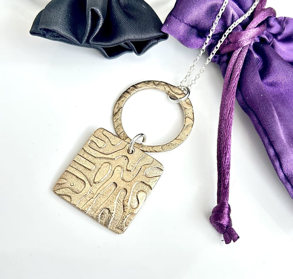 A handcrafted bronze clay double pendant, the bottom half is square with a textured surface, the top half is a bronze round ring with the same texture and they are linked together with a sterling silver jump ring. It has a sterling silver chain and is displayed on a white surface with two satin jewellery pouches, one purple and the other black.
