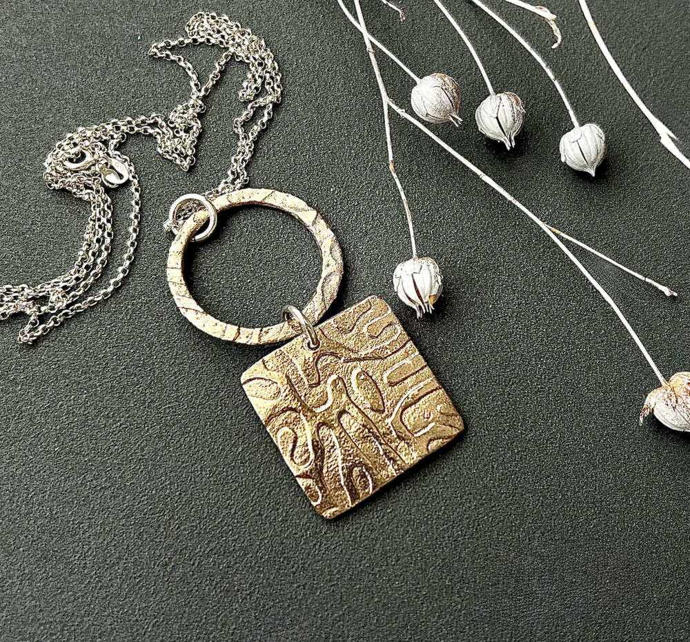 A handcrafted bronze clay double pendant, the bottom half is square with a textured surface, the top half is a bronze round ring with the same texture and they are linked together with a sterling silver jump ring. It has a sterling silver chain and is displayed on a dark grey surface.