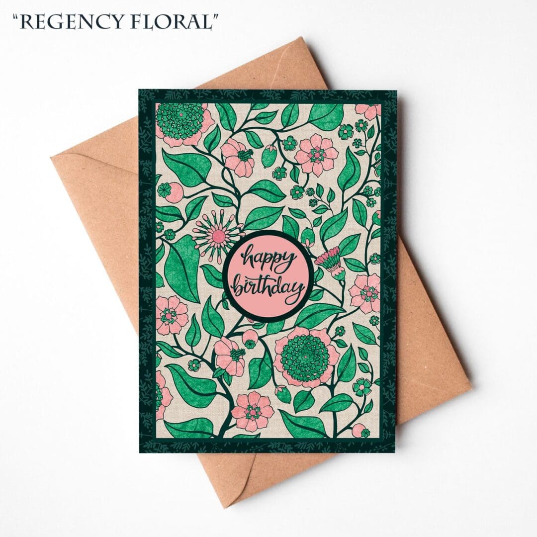 A hand-illustrated birthday card featuring elegant pink and coral Regency-style flowers with the words “Happy Birthday,” resting on a natural brown Kraft envelope.