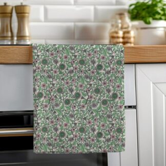 A green and pink floral cotton tea towel with a hand-illustrated Regency-inspired pattern, hanging neatly on a wooden oven rail in a bright kitchen.