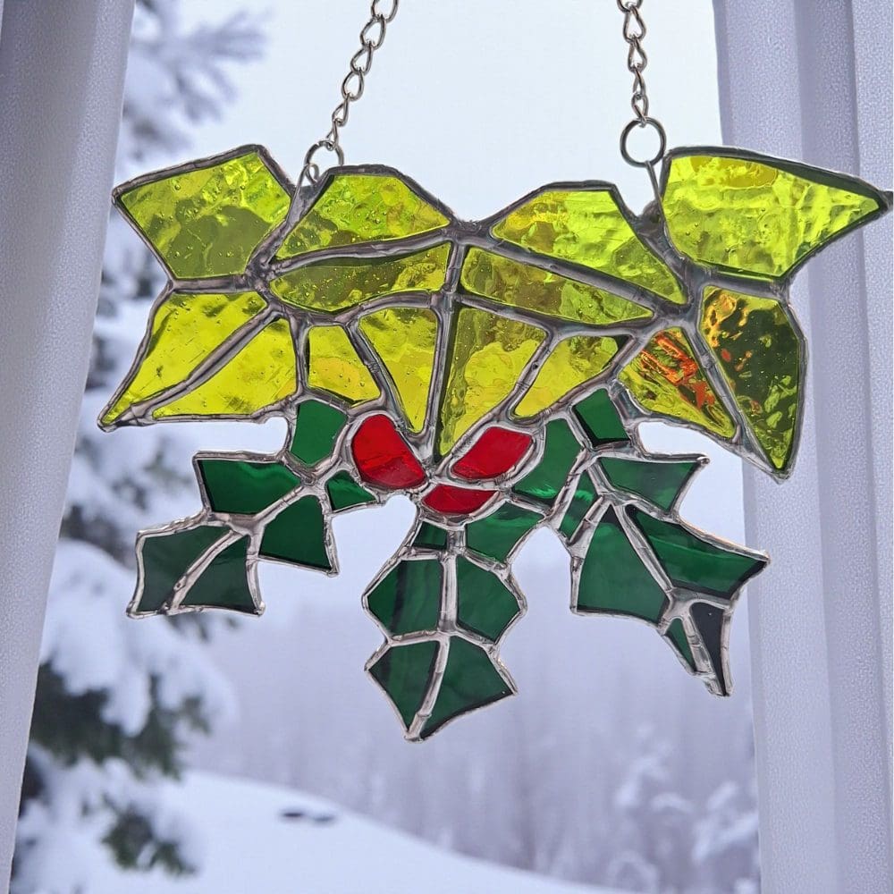 Handmade stained glass suncatcher featuring a wreath design of green holly leaves, ivy leaves, and vibrant red glass berries. Crafted using the copper foil technique in a UK studio from an original pattern. This small, unique piece of British-made luxury glass art is a festive, sentimental Christmas and winter window decoration and keepsake gift, hanging from a silver chain against a bright background.
