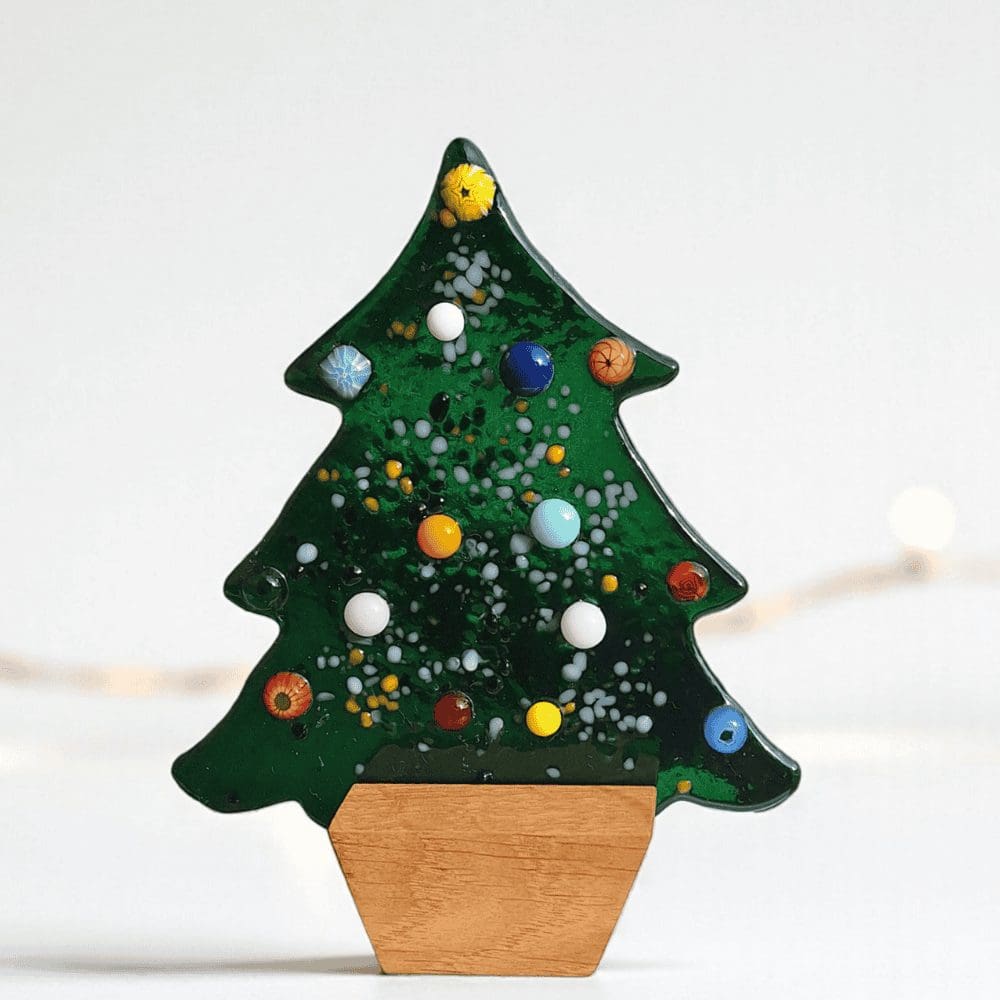 Small freestanding green fused glass Christmas tree with textured, colourful detail, resting in a simple square wooden base. This handmade, unique glass decoration is 13.5cm tall and is perfect for a mantel or shelf display. It is sold as a luxury holiday gift or home accessory. It is sent in two pieces, wrapped in tissue paper and purple ribbon, ready to be given as a present.