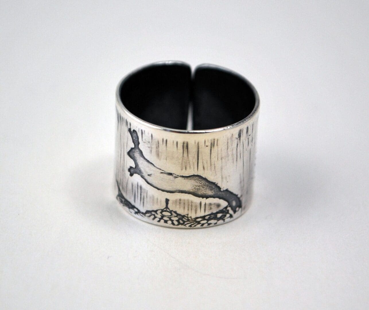 A side view of a wide, sterling silver cuff ring, showing the open gap and an etched, oxidized design of a leaping hare on a brushed metal background