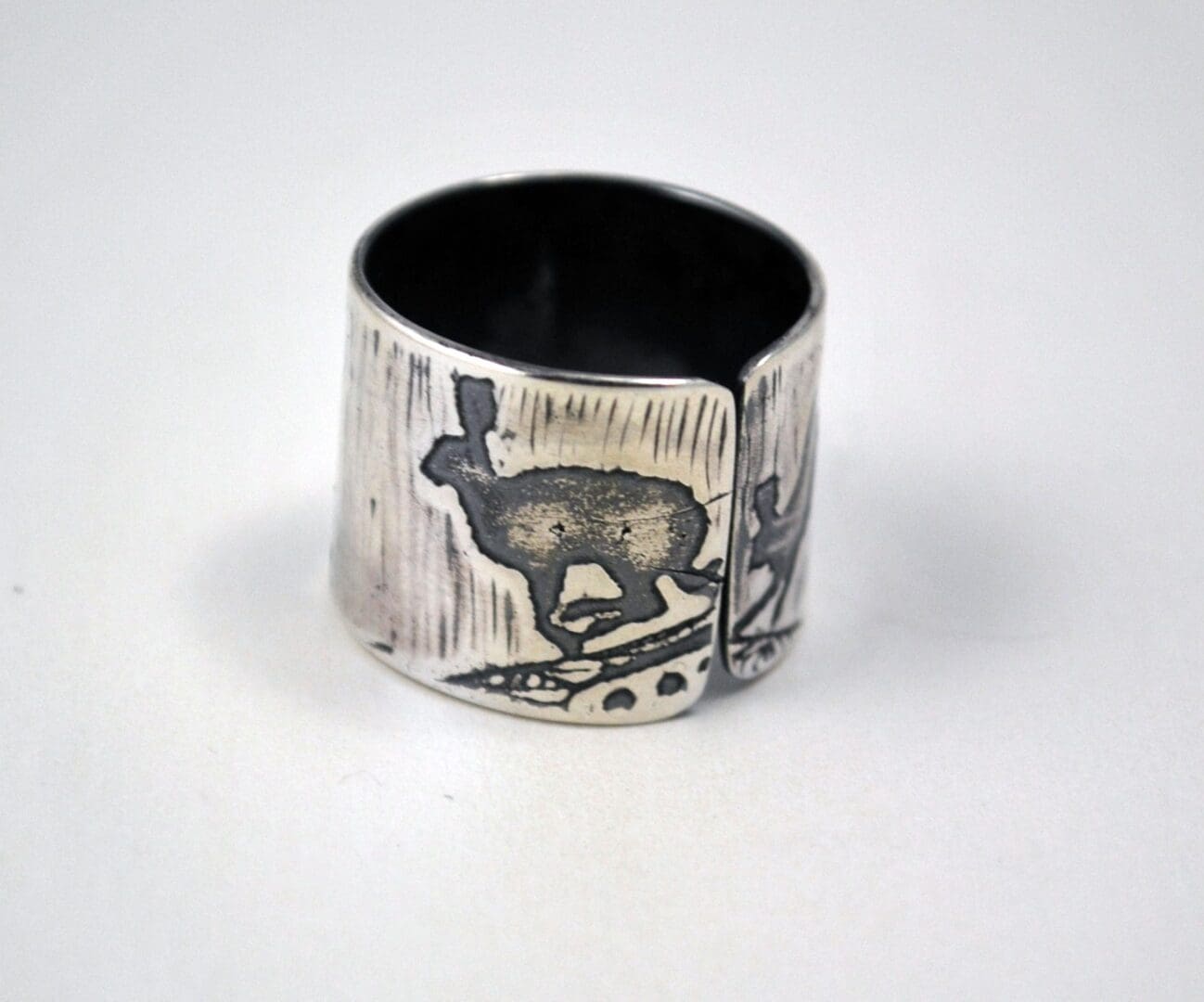 A close-up, slightly angled view of a wide, sterling silver cuff ring with an etched and oxidized design of a running hare, showing the open gap and brushed texture
