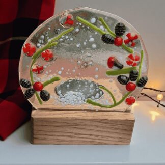 Handmade fused glass tealight holder featuring a robin surrounded by holly leaves and red berries. The circular glass panel measures 9.5 cm across and 8.5 cm high and sits in a wooden base 8 cm wide and deep. The total height is approximately 10 cm. Designed for a tealight candle (battery-operated recommended), it glows beautifully when lit. Presented wrapped in white tissue paper with a purple ribbon, it can be sent directly as a handwritten gift.