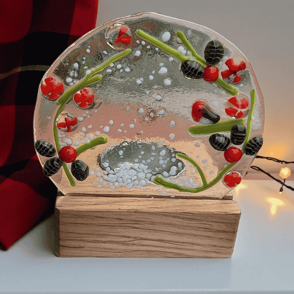 Handmade fused glass tealight holder featuring a robin surrounded by holly leaves and red berries. The circular glass panel measures 9.5 cm across and 8.5 cm high and sits in a wooden base 8 cm wide and deep. The total height is approximately 10 cm. Designed for a tealight candle (battery-operated recommended), it glows beautifully when lit. Presented wrapped in white tissue paper with a purple ribbon, it can be sent directly as a handwritten gift.