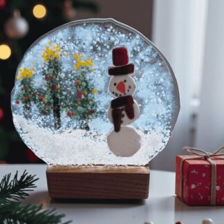 Handmade fused glass snowman decoration with a wooden candle holder base. The clear glass features a cheerful snowman wearing a red hat and scarf, surrounded by snowy trees and falling snow. A warm winter light shines through the glass, highlighting the textures and colours of the fused design. This artisan glass ornament makes a charming handmade Christmas decoration or gift, lovingly wrapped in white tissue paper with a purple ribbon, ready for gifting.